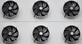 Six industrial air conditioning condenser fans mounted on an exterior facility wall for large-scale cooling