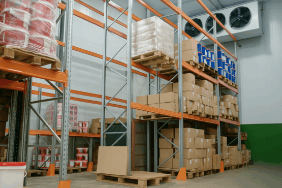 Warehouse interior showing tall racking shelves stocked with boxes and paint buckets, featuring wall-mounted industrial cooling fans for cold storage.