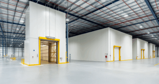 Wide-angle view of an industrial warehouse with insulated cold storage rooms, yellow-framed access doors, and a high ceiling with steel beams and lighting.
